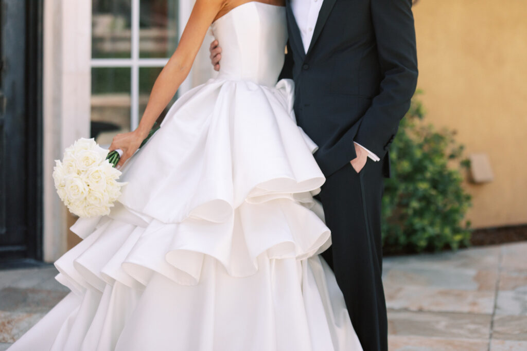 A bride in a strapless white wedding dress holding a bouquet of white flowers stands next to a groom in a black suit, posing outdoors at an elegant event organized by a Northern California wedding planner.