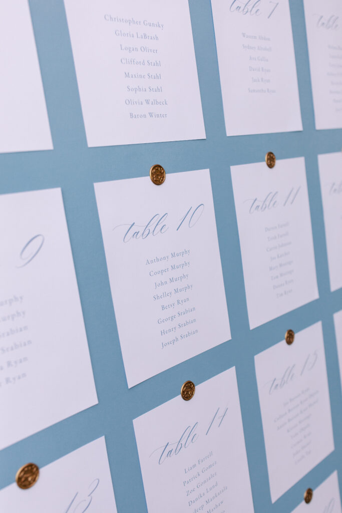 A blue board displays a grid of white cards with table assignments and guest names, each card pinned with a gold seal.