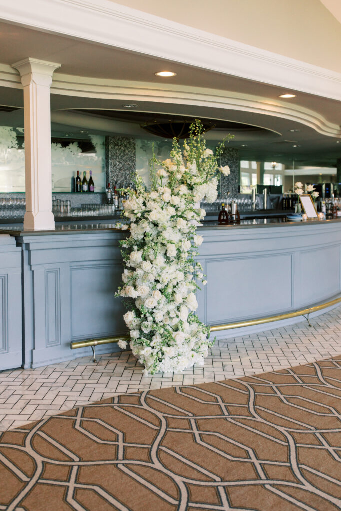 A tall vertical arrangement of white flowers with greenery stands in front of a gray bar counter in a spacious, well-lit room with patterned carpet and tiled floor.