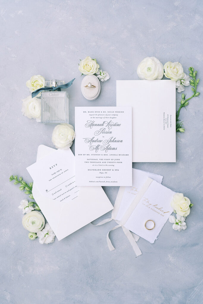 Flat lay of elegant wedding invitations, RSVP card, two vow books, a ring box with an engagement ring, a perfume bottle, and white flowers on a light blue textured surface.