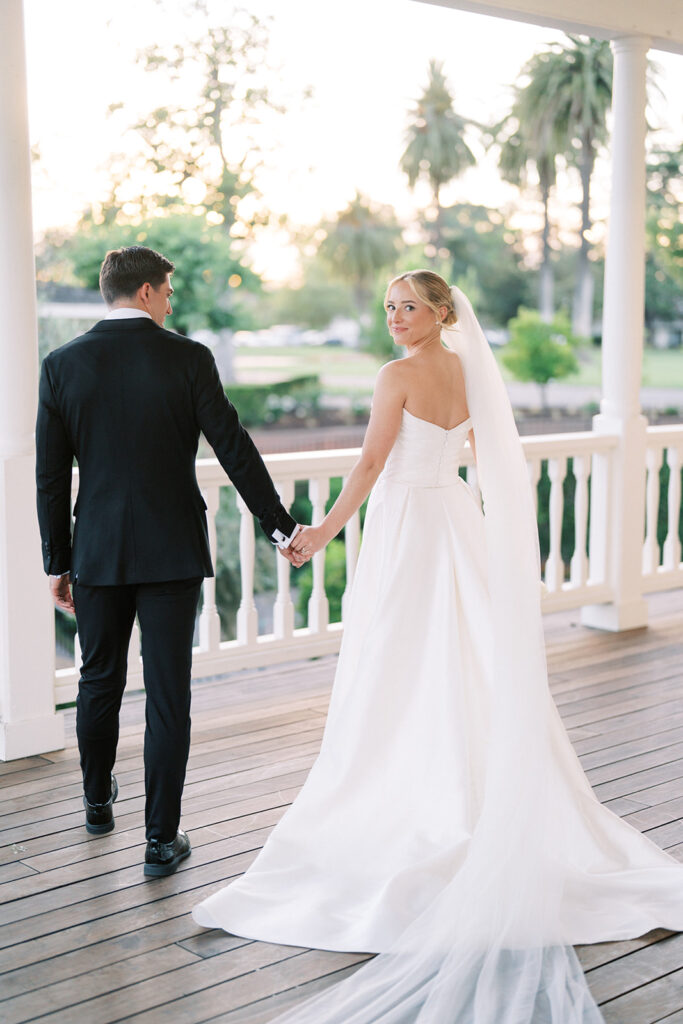 A bride in a white gown and veil holds hands with a groom in a black suit as they walk on a wooden porch with white railings, surrounded by greenery.