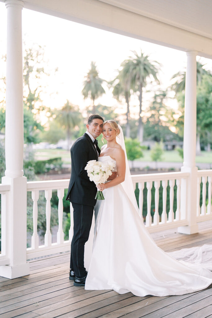 A bride and groom stand together on a porch, the bride holding a bouquet of white flowers and wearing a long white gown and veil, with greenery and palm trees in the background.