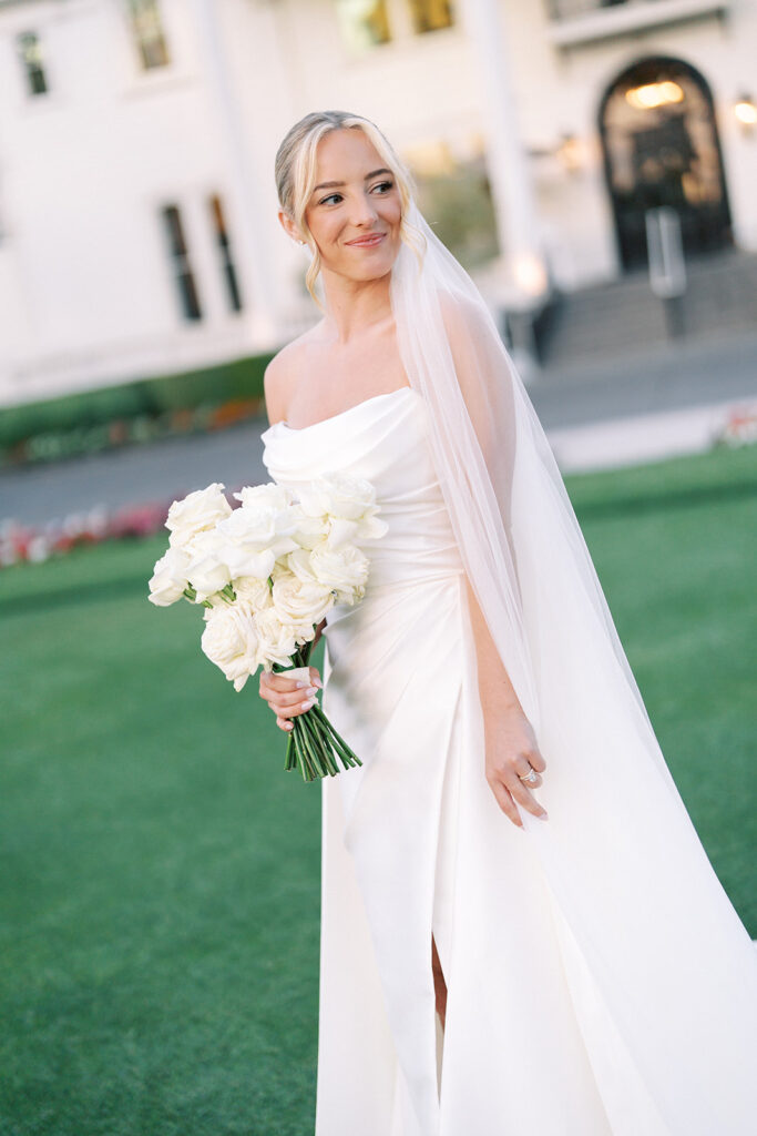 A bride in a white dress and veil stands outdoors holding a bouquet of white roses, with a building in the background.