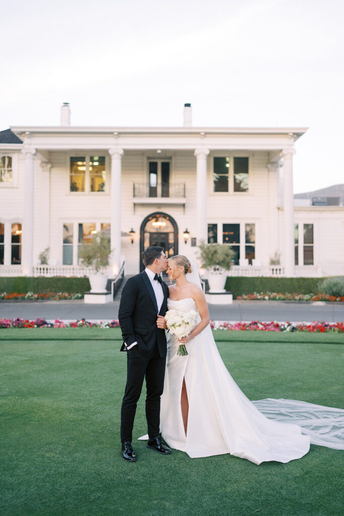 A bride and groom stand together on a lawn in front of a large white building, with the bride holding a bouquet and wearing a strapless gown with a train.