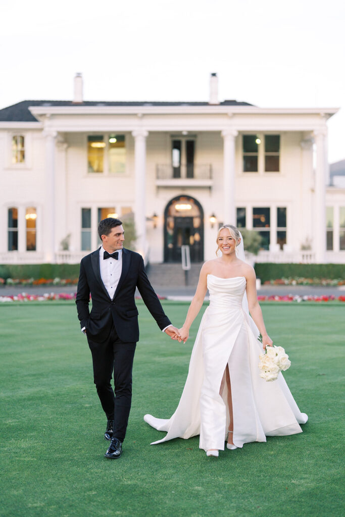 A bride and groom in formal attire walk hand in hand on a lawn in front of a large white house.