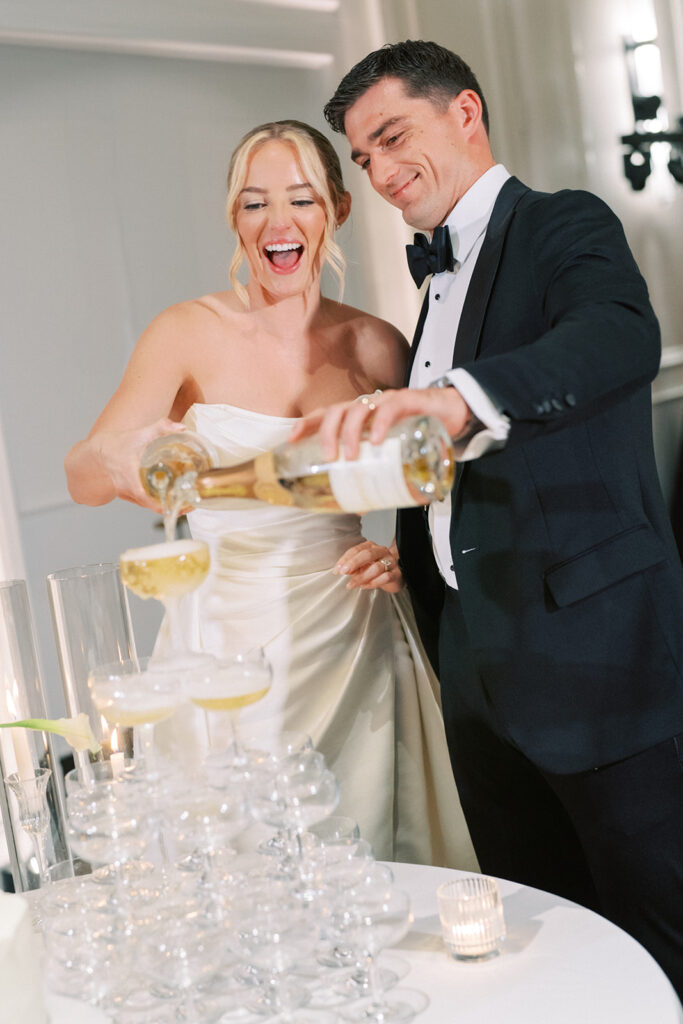A bride and groom in formal attire pour champagne together into a tiered tower of glasses at a wedding reception.