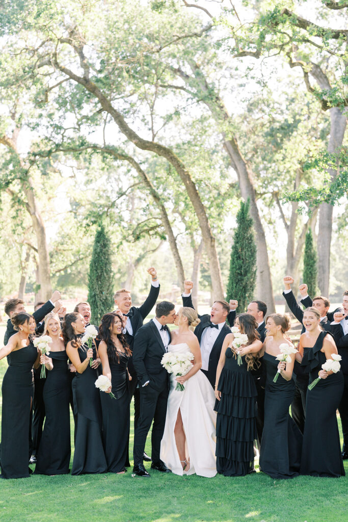 A bride and groom kiss outdoors while their wedding party, dressed in black formal attire, stand around them holding white bouquets and cheering. Tall trees and greenery are in the background.