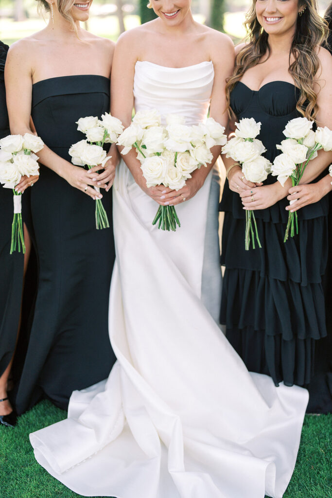 Three women in formal dresses holding white rose bouquets; the woman in the center wears a white gown, while the others wear black dresses.