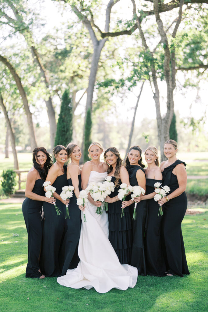 A bride in a white dress stands outdoors with seven bridesmaids in black dresses, all holding white bouquets, with trees and grass in the background.