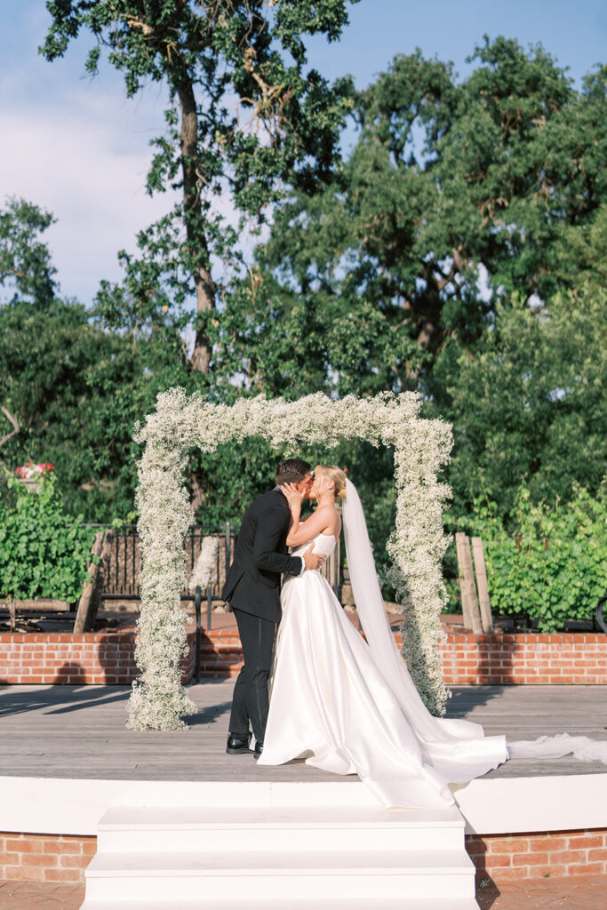 A bride and groom kiss under a white floral arch during an outdoor wedding ceremony, surrounded by greenery and trees.