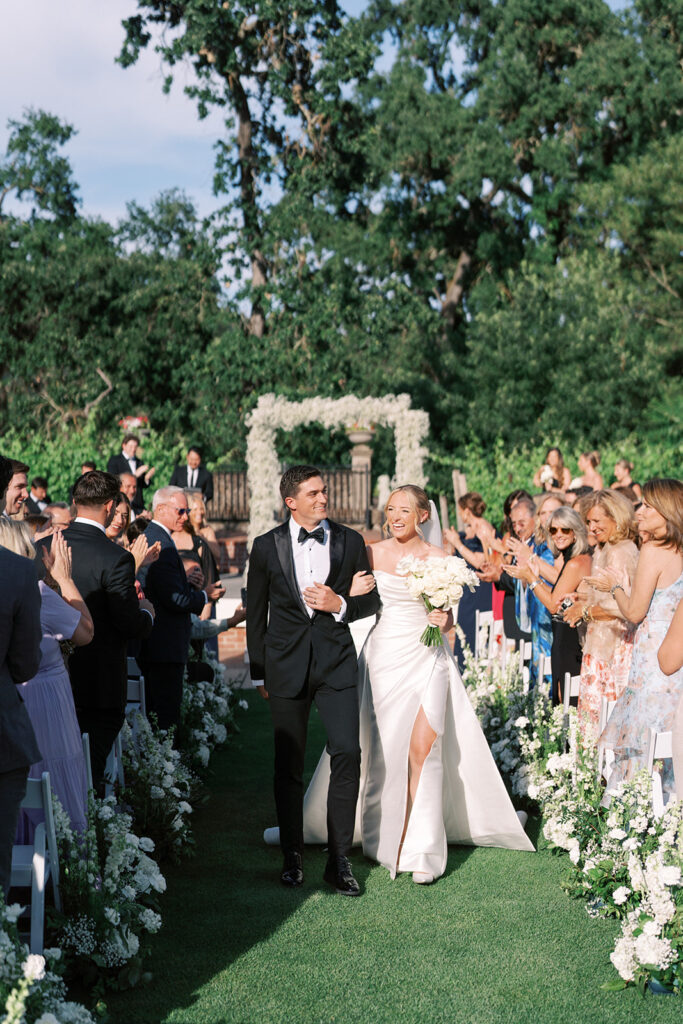 A bride and groom walk down an outdoor aisle lined with white flowers as guests stand and applaud.