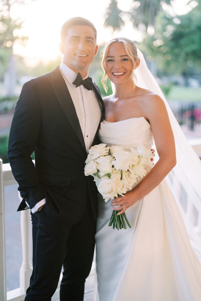 A bride in a white gown holds a bouquet of white roses and stands next to a groom in a black tuxedo, both smiling at the camera outdoors.