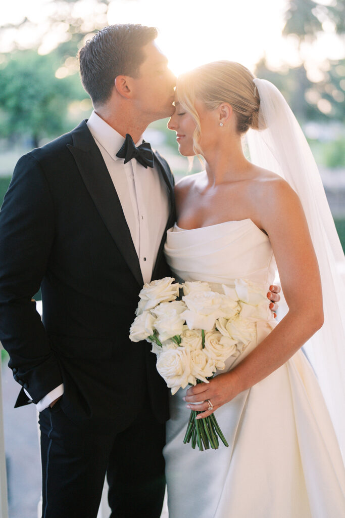 A groom in a black tuxedo kisses the forehead of a bride in a white gown holding a bouquet of white roses, both standing closely together outdoors.