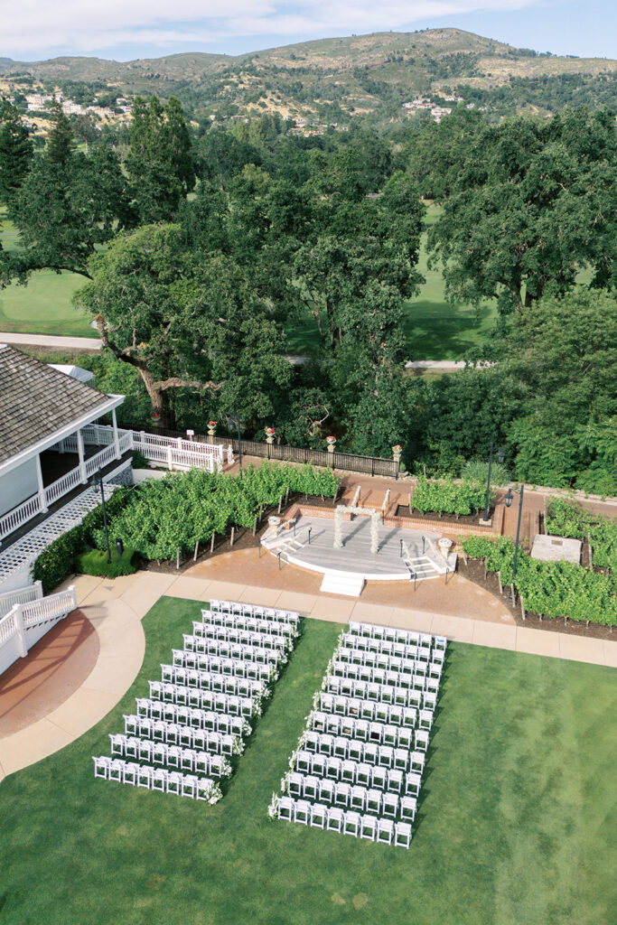 Rows of white chairs are arranged on a lawn facing a small outdoor stage, with trees and hills in the background.