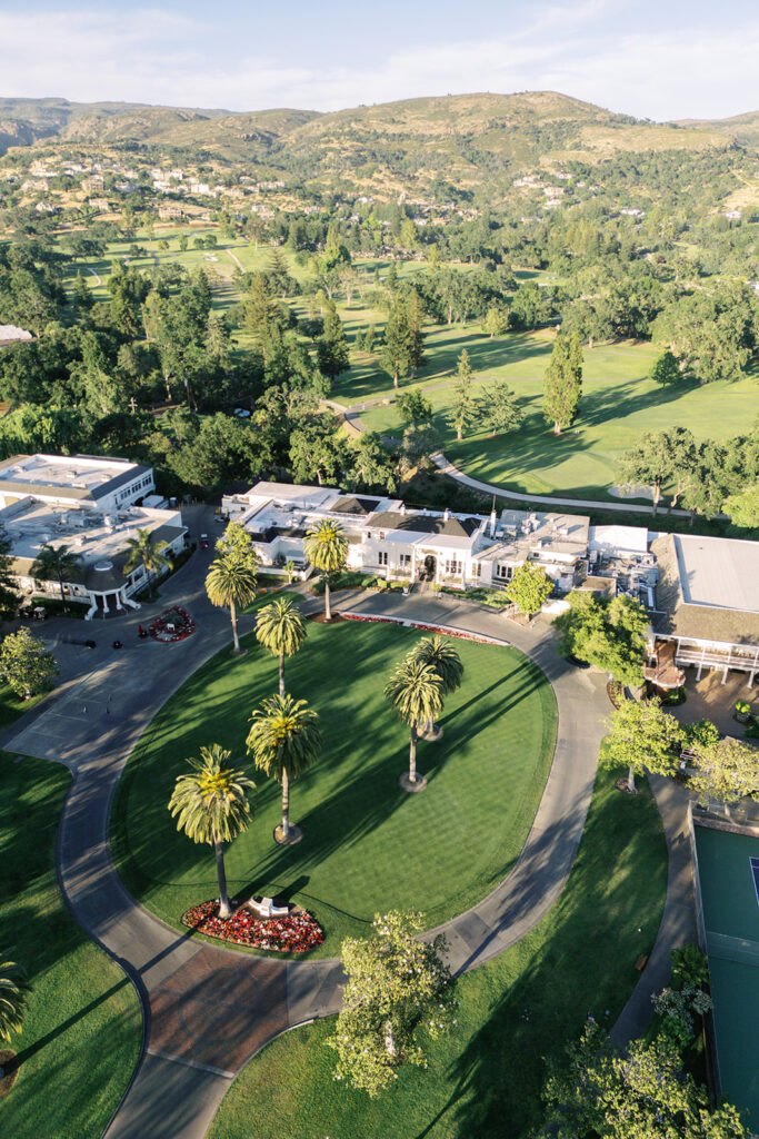 Aerial view of a circular driveway with palm trees, surrounded by buildings and a golf course in a hilly, green landscape.