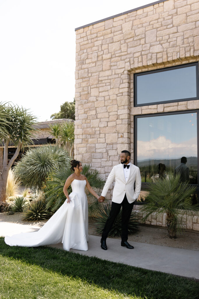 A bride in a white dress and a groom in a white jacket and black pants hold hands outside a stone building with large windows and desert plants.