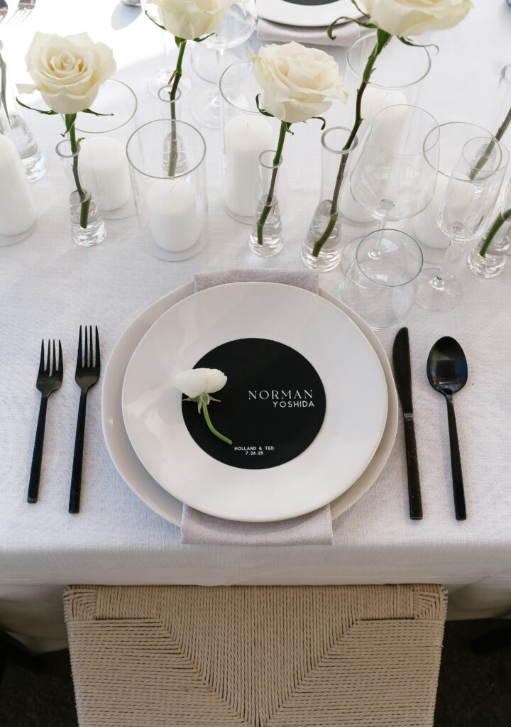 A formal table setting with a white plate featuring a black card labeled "Norman Yoshida," black cutlery, glassware, white roses, and candles on a white tablecloth.