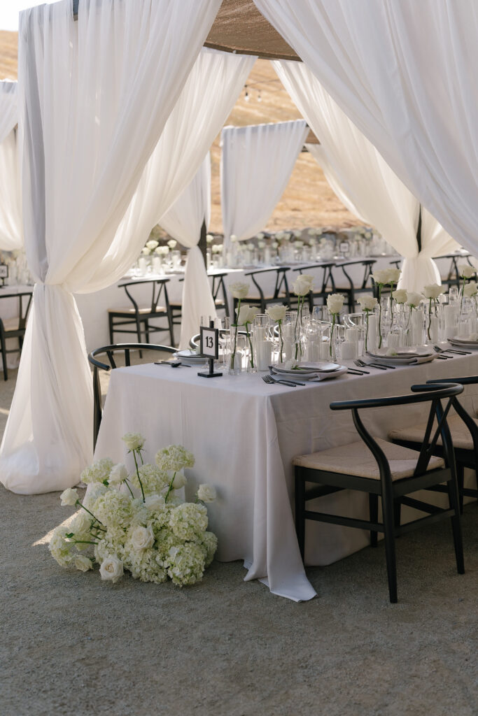 A table set for an event with white tablecloth, glassware, black chairs, and white curtains, with floral arrangements and table number 13 visible.
