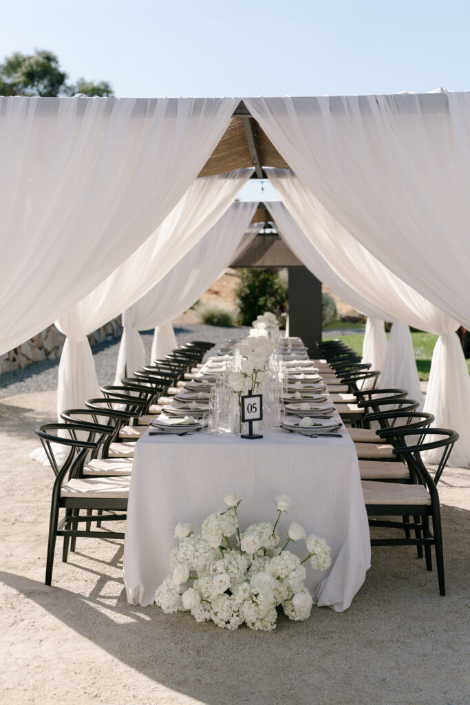 An outdoor dining table is set under a white draped canopy, with black chairs, clear glassware, white plates, and a centerpiece of white flowers. A table number card reads "05.