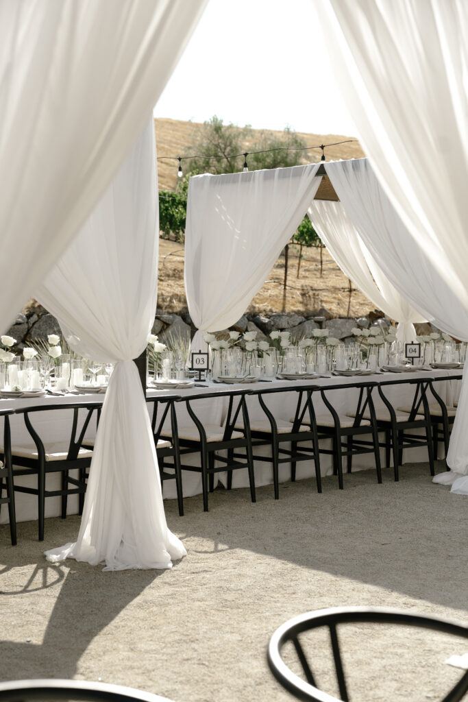 A long dining table with black chairs is set under white draped fabric outdoors, with glassware and white flowers arranged on the table.