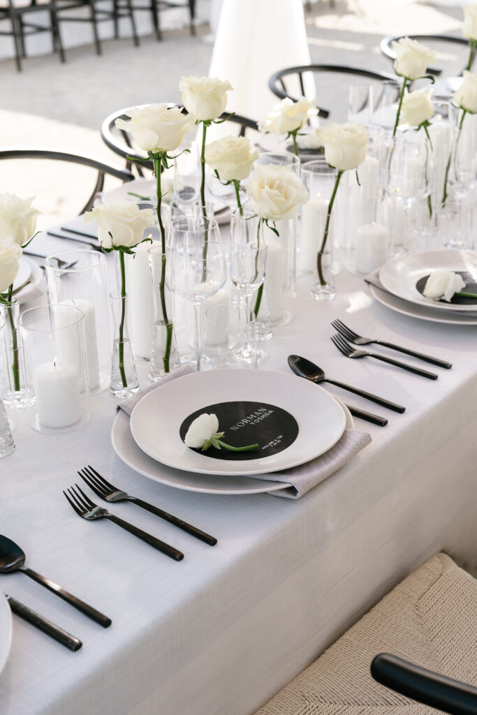 A formal table setting with white plates, black cutlery, black menus, white napkins, and tall white roses in glass vases on a white tablecloth.