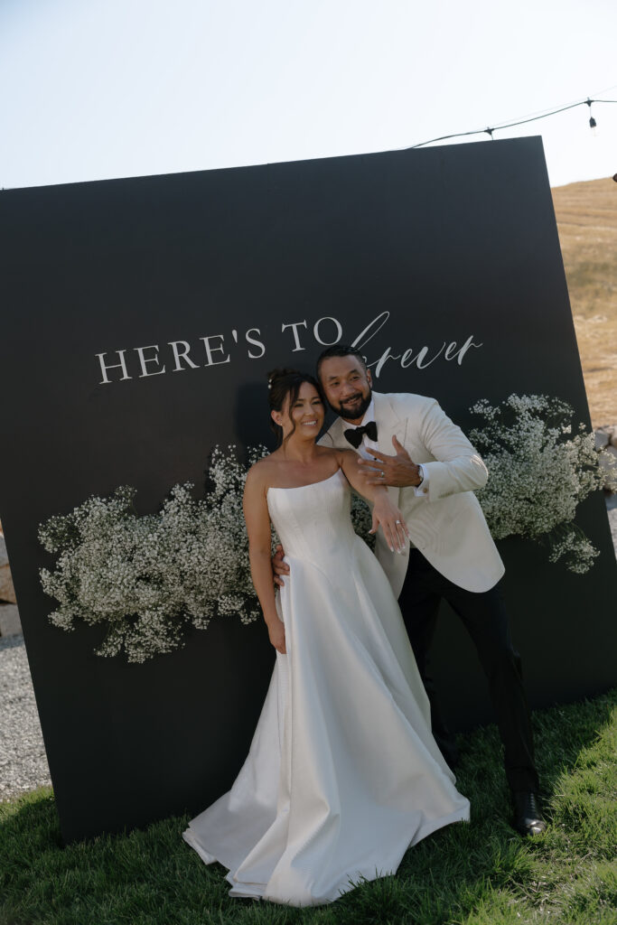 A bride and groom pose and smile together in front of a black backdrop that reads "Here's to forever," decorated with white flowers.