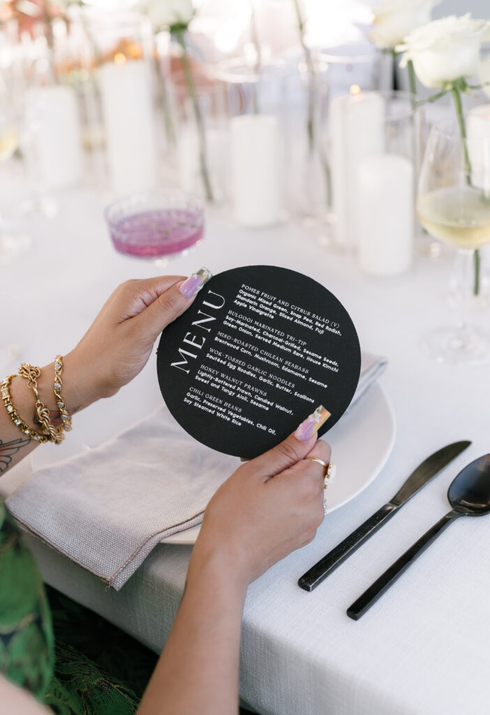 A person holding a round black menu at a formally set dining table with candles, flowers, glassware, and cutlery.