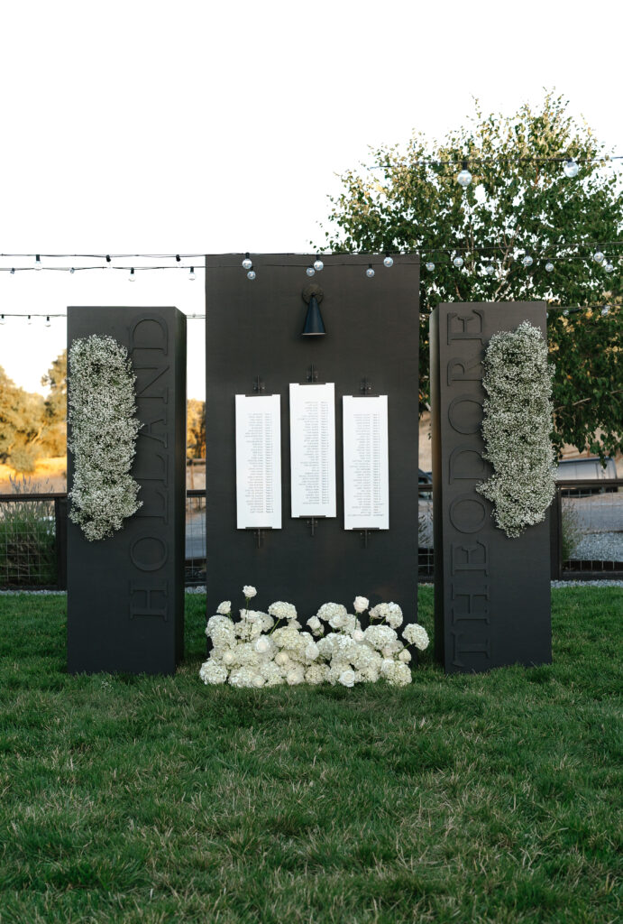 Three black display boards with white flowers and hanging white papers stand on grass; the boards read "Holland" and "Theodore" with flowers at the base and a tree in the background.