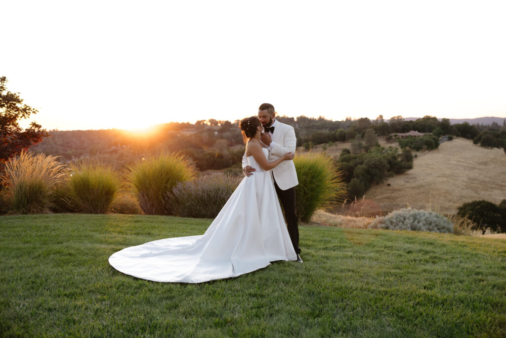 A bride and groom embrace on a grassy hill at sunset, with rolling hills and trees in the background.