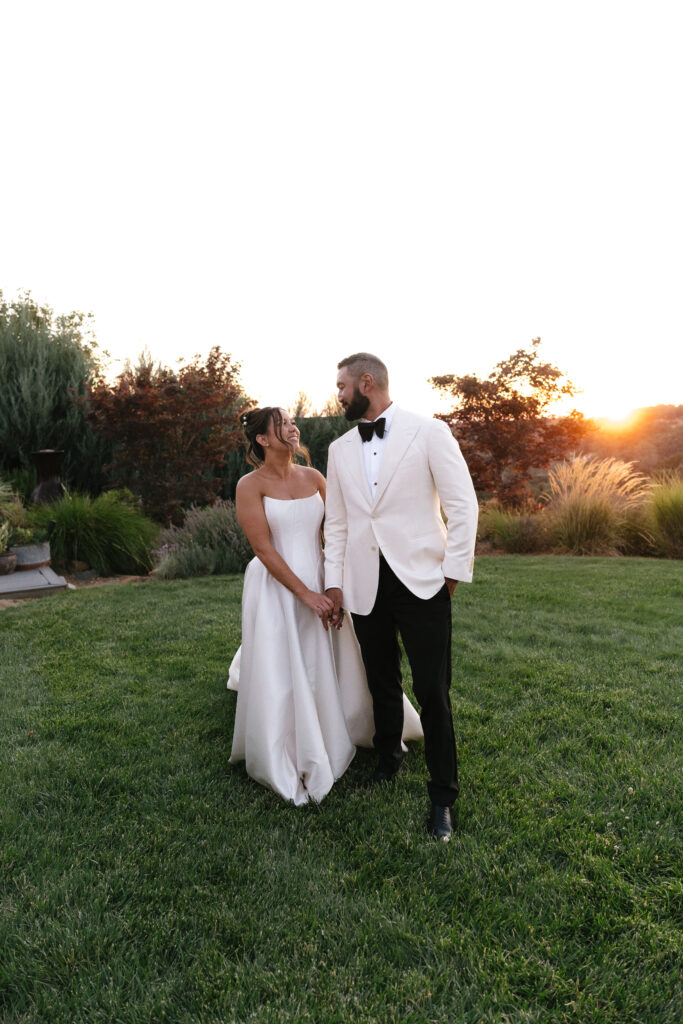 A bride and groom stand together on a grassy lawn at sunset, holding hands and looking at each other, with trees and shrubs in the background.