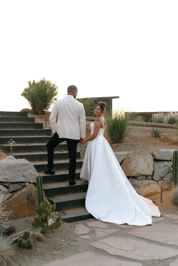 A bride in a white gown and a groom in a white jacket walk up outdoor stone steps, holding hands, surrounded by desert plants and rocks.