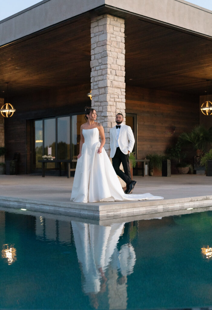 A bride in a white gown and a groom in a white jacket stand by the edge of a modern building near a pool, with their reflections visible in the water.