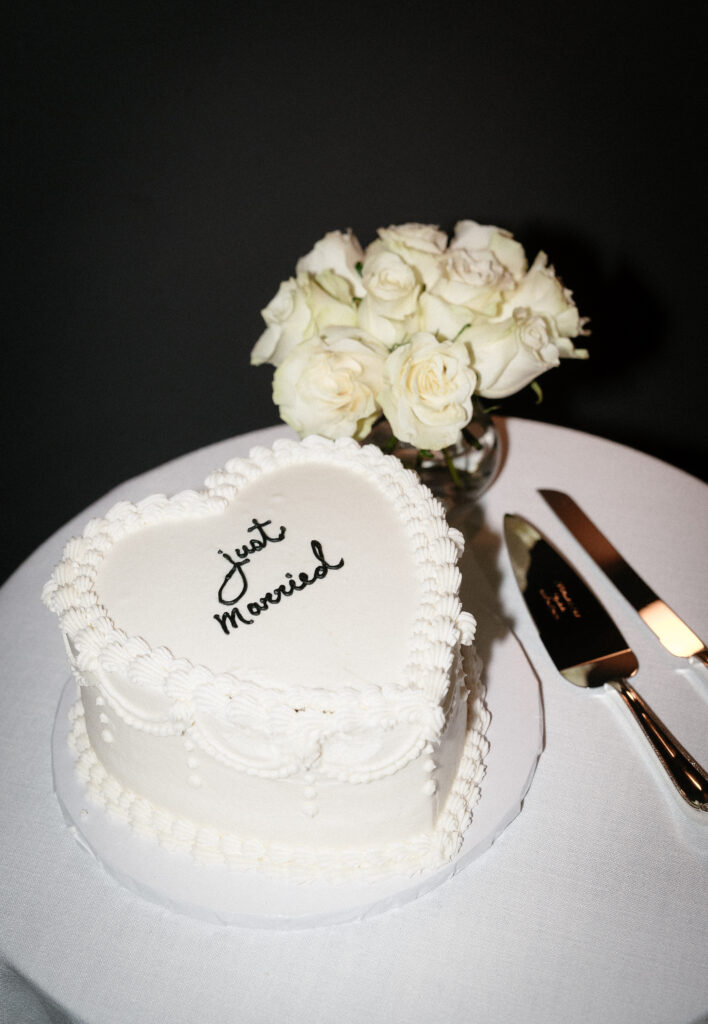 A heart-shaped white cake with "Just married" written on top sits on a table next to a bouquet of white roses and a cake knife and server.