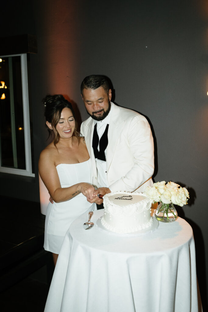 A couple stands together cutting a white cake on a round table with a vase of white flowers in a dimly lit room.