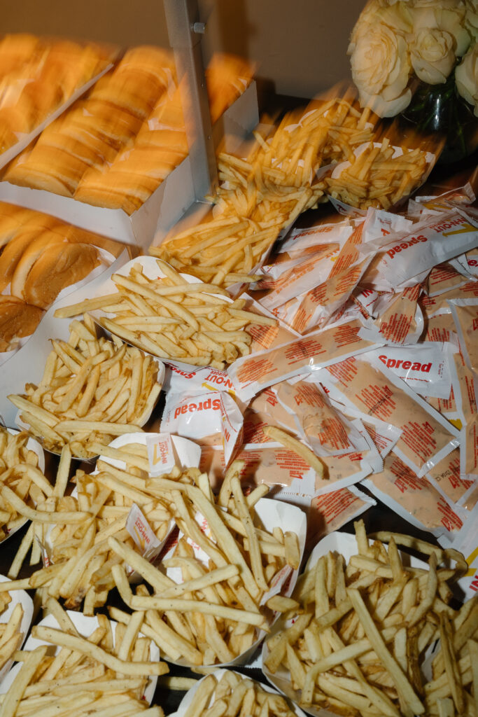 Several servings of French fries in trays, unopened ketchup packets, and buns arranged on a table, with a bouquet of white roses in the corner.