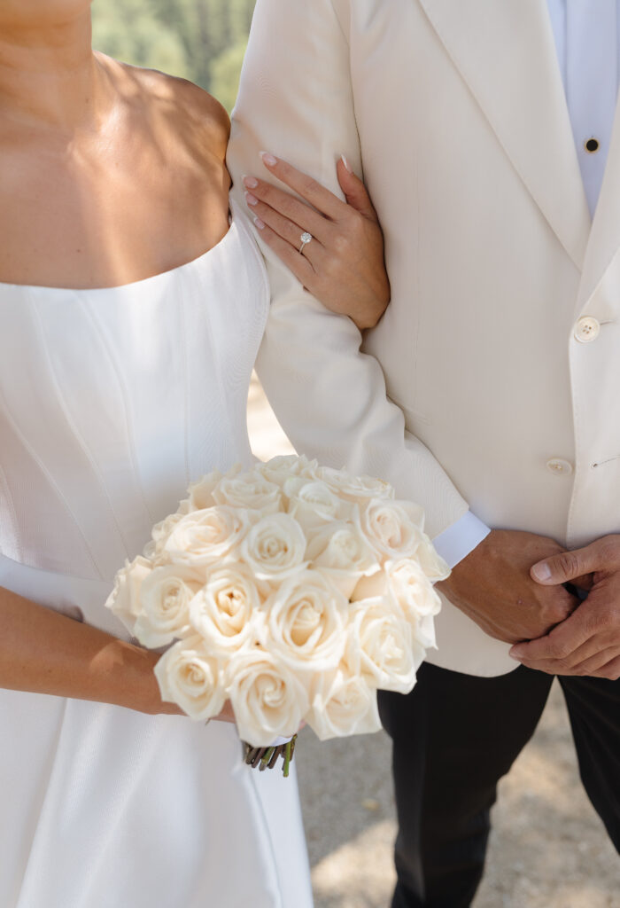 A bride in a white dress holds a bouquet of white roses and links arms with a groom in a cream-colored suit. Her engagement ring is visible.