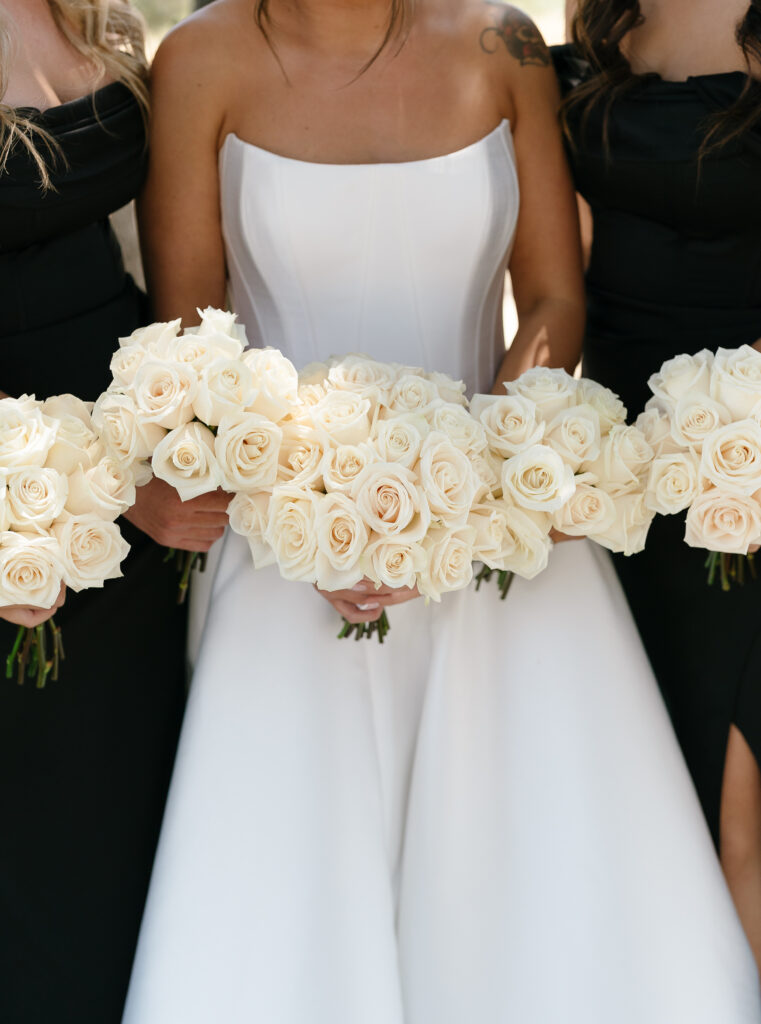 Three women in formal dresses hold bouquets of white roses; the woman in the center wears a white strapless gown, while the others wear black.