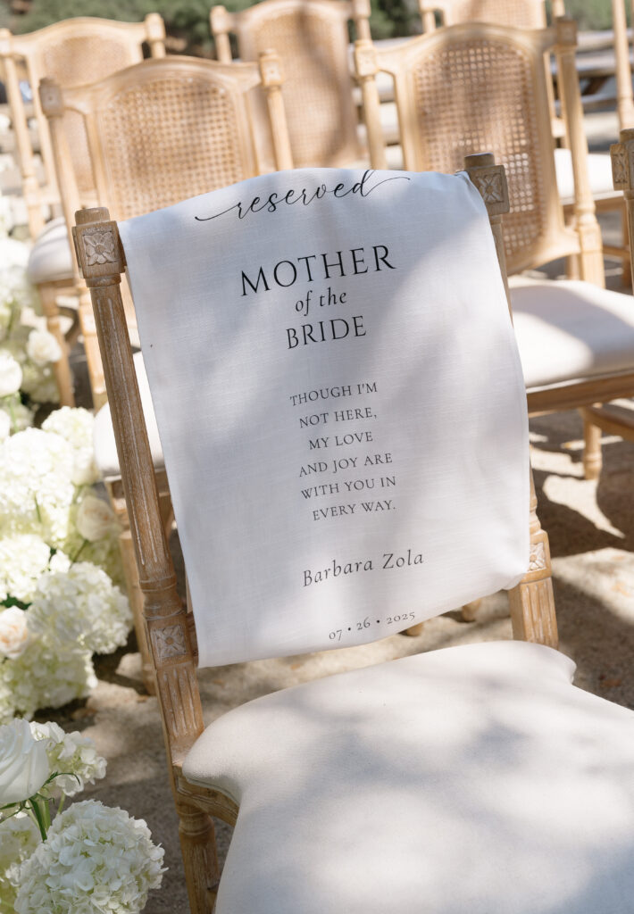 A wooden chair with a white cloth reading "reserved Mother of the Bride," with a memorial message and the name Barbara Zola, at an outdoor wedding ceremony.