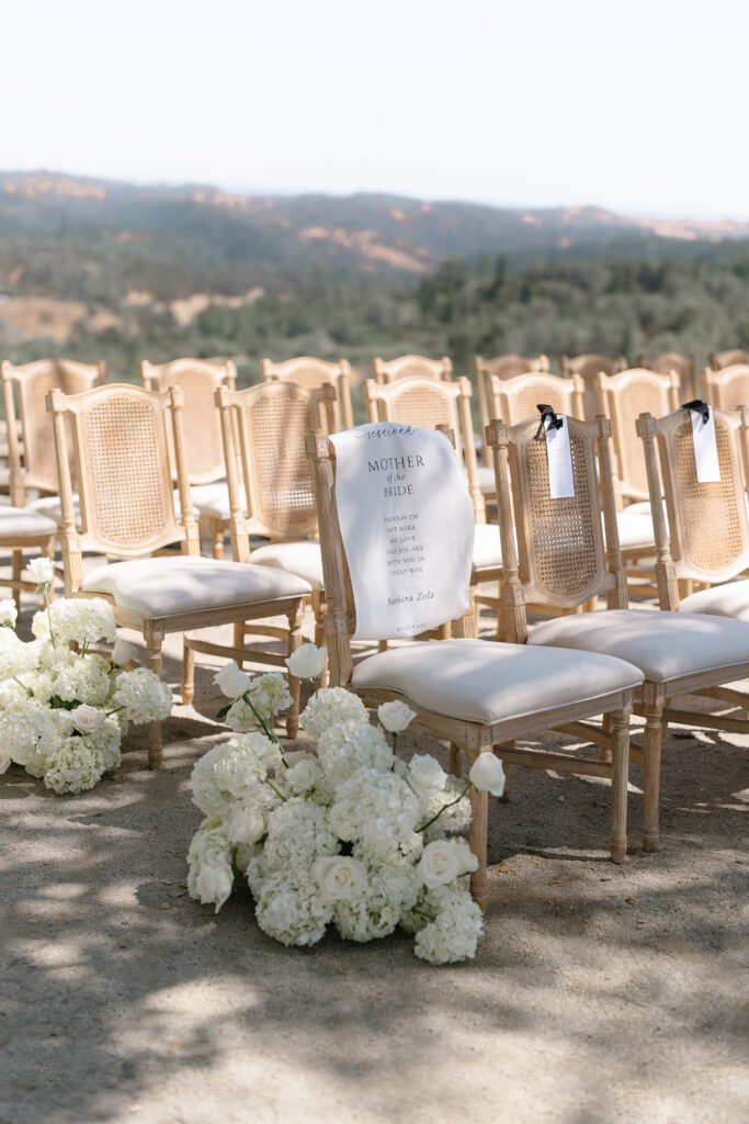 Rows of wooden chairs with white cushions set outdoors; one chair reserved with a sign and white floral arrangements placed in front.