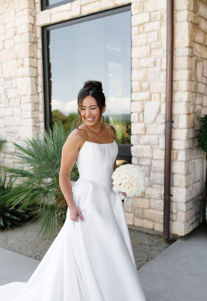 A woman in a white off-the-shoulder wedding dress holding a bouquet of white roses stands in front of a stone building and smiles.