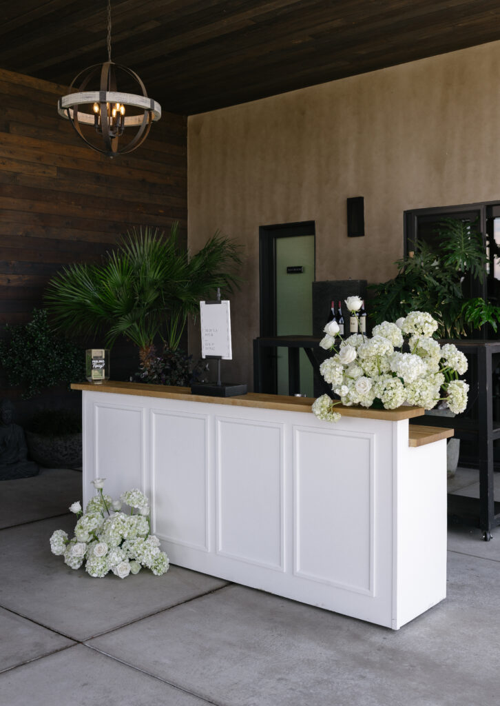 A white reception desk with floral arrangements and a sign stands in a modern entryway with greenery and dark wood accents.