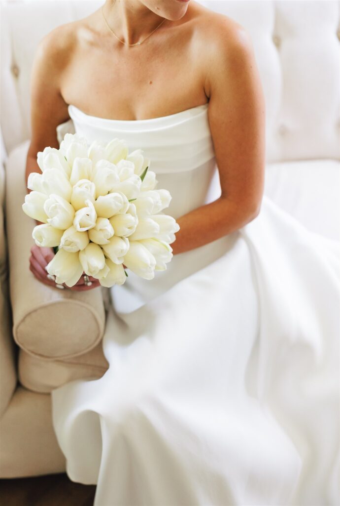 A woman in a white strapless dress sits on a beige couch, holding a bouquet of white tulips in her lap.