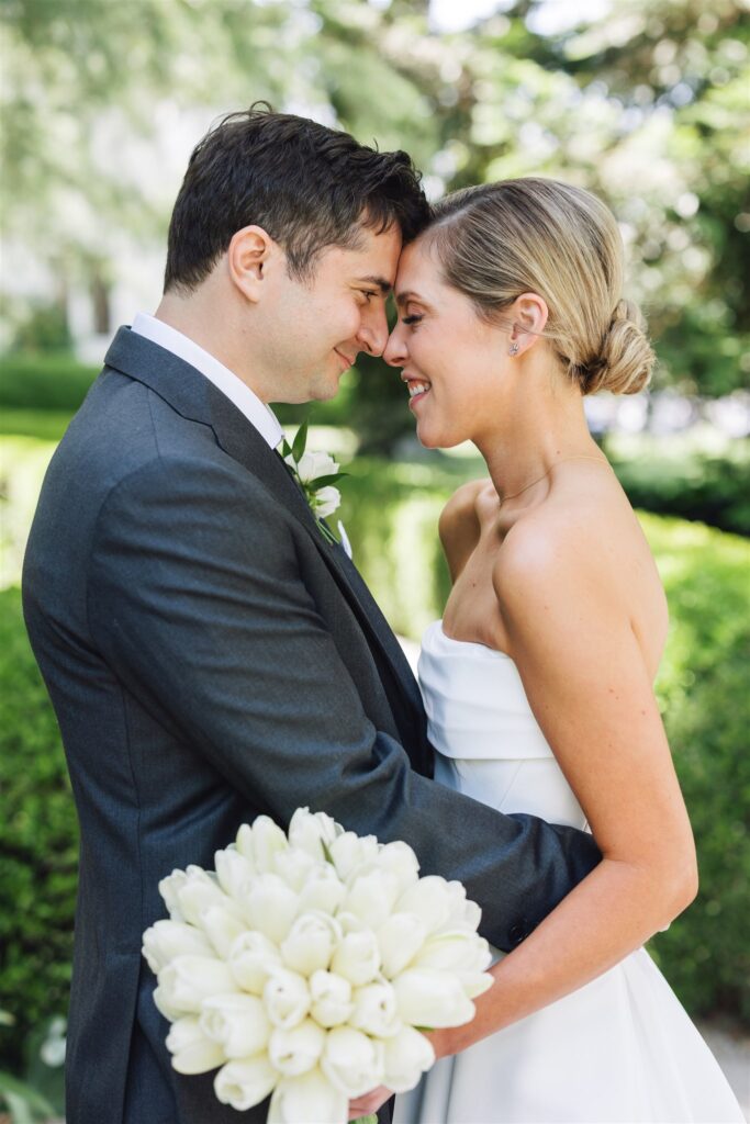 A bride and groom stand close together outdoors, smiling with foreheads touching. The bride holds a bouquet of white flowers.