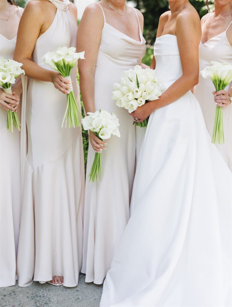Four women in light-colored dresses, one in a white wedding gown, each holding a bouquet of white flowers, standing side by side.