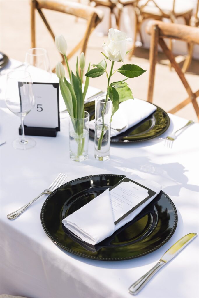A table setting with a black plate, folded white napkin, menu card, and glass vases holding white tulips and a rose on a white tablecloth.