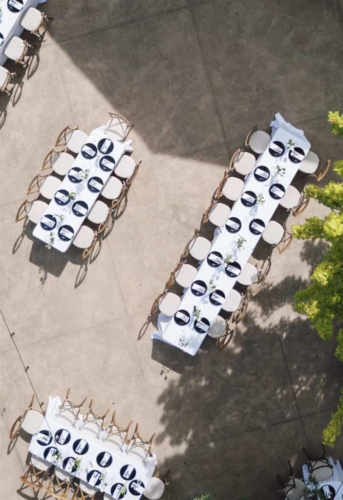 Aerial view of two long banquet tables with white tablecloths and place settings, arranged outdoors on a concrete surface with some nearby greenery.