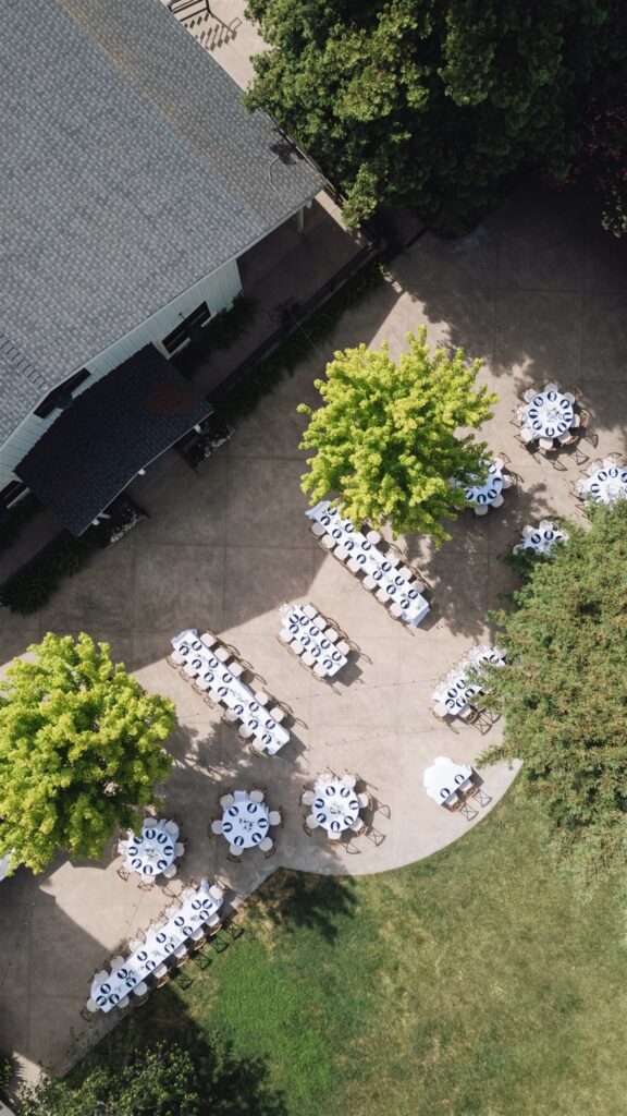 Aerial view of outdoor event setup with round and rectangular tables covered in white tablecloths, arranged on a paved area near trees and a building.