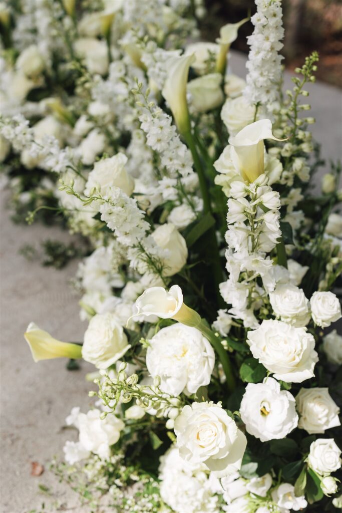 Arrangement of white flowers, including roses, calla lilies, and delphiniums, placed on a light surface.