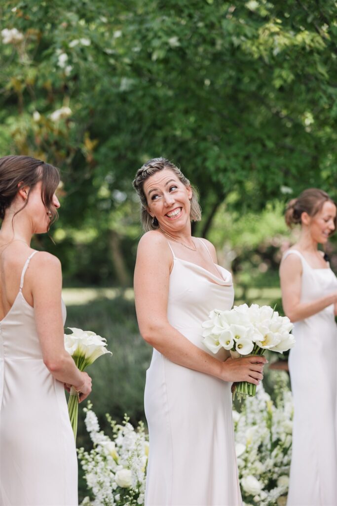 Three women in white dresses stand outdoors holding bouquets of white flowers, with greenery and white floral arrangements in the background.