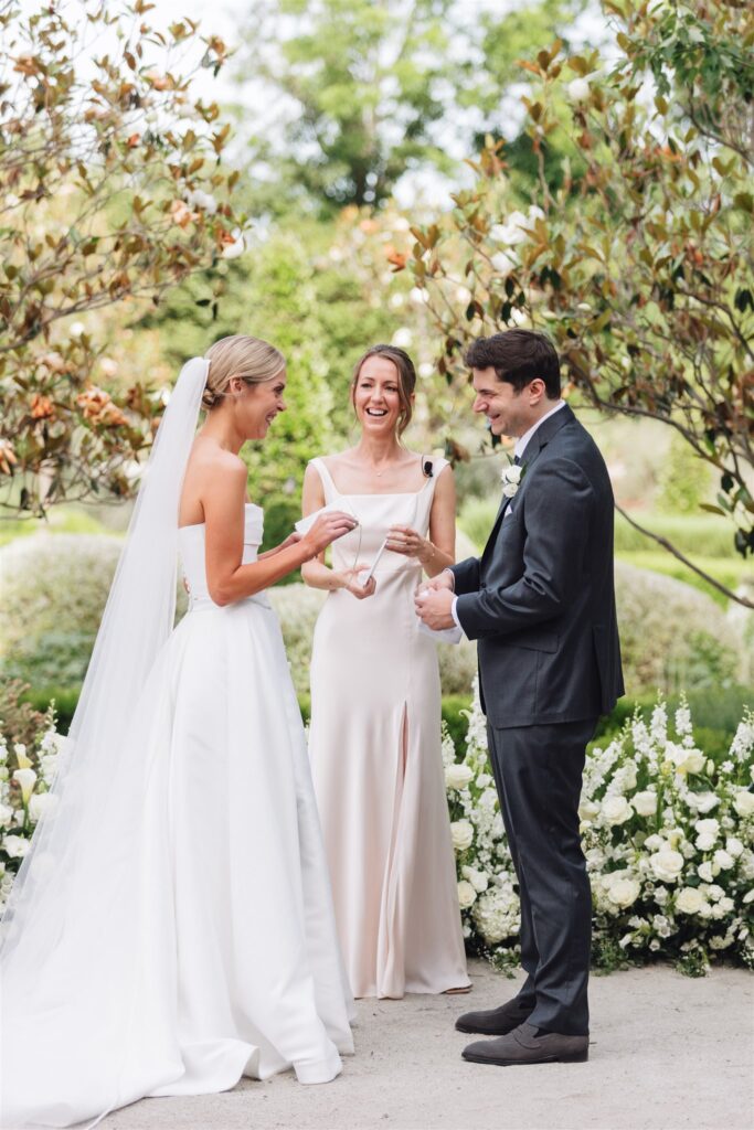 A bride and groom stand outdoors exchanging vows, smiling, with an officiant between them and white flowers in the background.